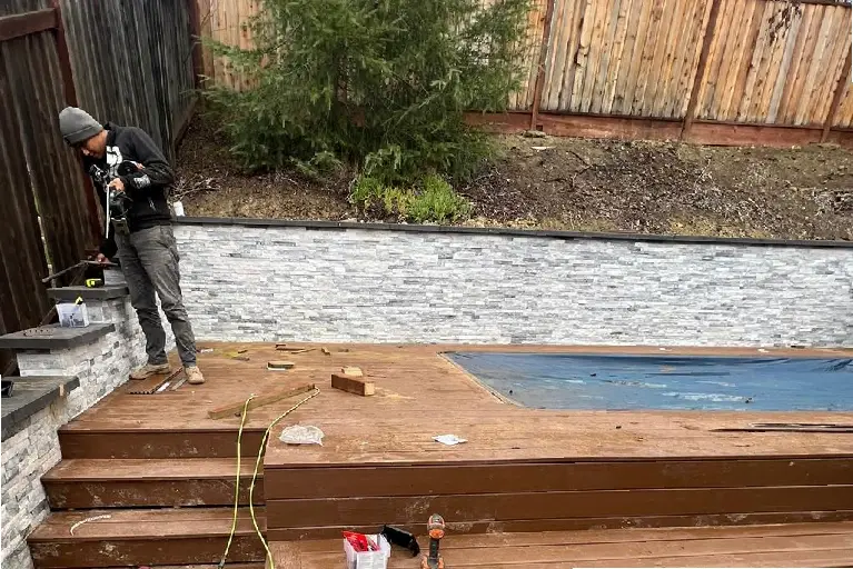 Hardscape contractor in Irvine building a modern stone retaining wall and composite deck near a backyard pool under construction.