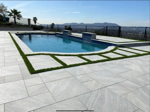 Elegant hilltop backyard featuring a custom pool with raised spillover fountains, surrounded by modern porcelain pavers and green turf accents—an eye-catching hardscape design by Papaya Construction in Irvine, CA, perfect for luxury outdoor living with breathtaking views.