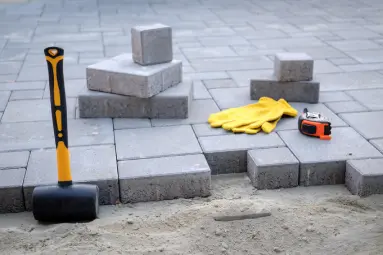 Paver installation in Huntington Beach showing rubber mallet, gloves, tape measure, and concrete blocks arranged on a compacted sand base for a patio project.