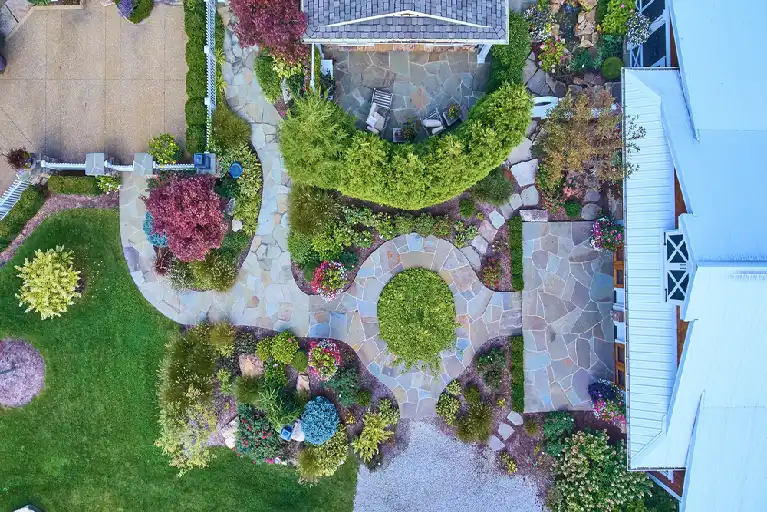 Aerial view of a beautifully landscaped residential backyard featuring curved stone pathways, lush greenery, flower beds, and a circular patio area surrounded by trees and shrubs next to a white-roofed house.