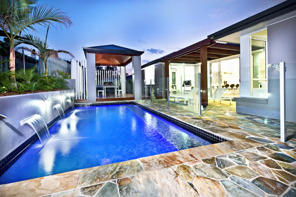 Contemporary backyard with a sleek pool, cascading water features, glass fencing, and elegant stone patio in a luxury outdoor living space