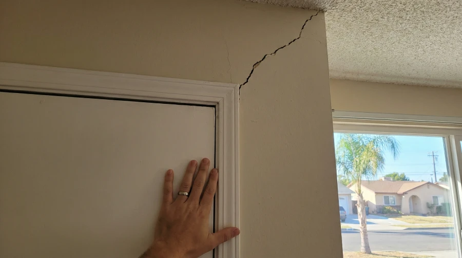 A homeowner's hand pushing against a sticking interior door beneath a large diagonal wall crack, a major warning sign of foundation problems in a Santa Ana, CA home.