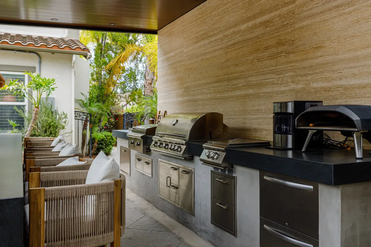 Built-in stainless steel grills on a granite countertop in front of a travertine wall in a Yorba Linda outdoor kitchen