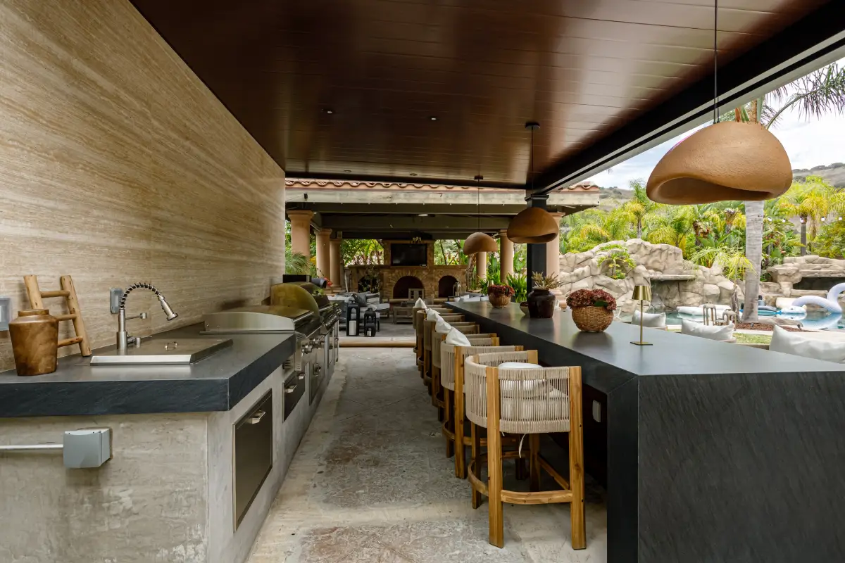 Covered outdoor kitchen in Yorba Linda with a sink, granite countertops, a travertine wall and a long black bar with stools